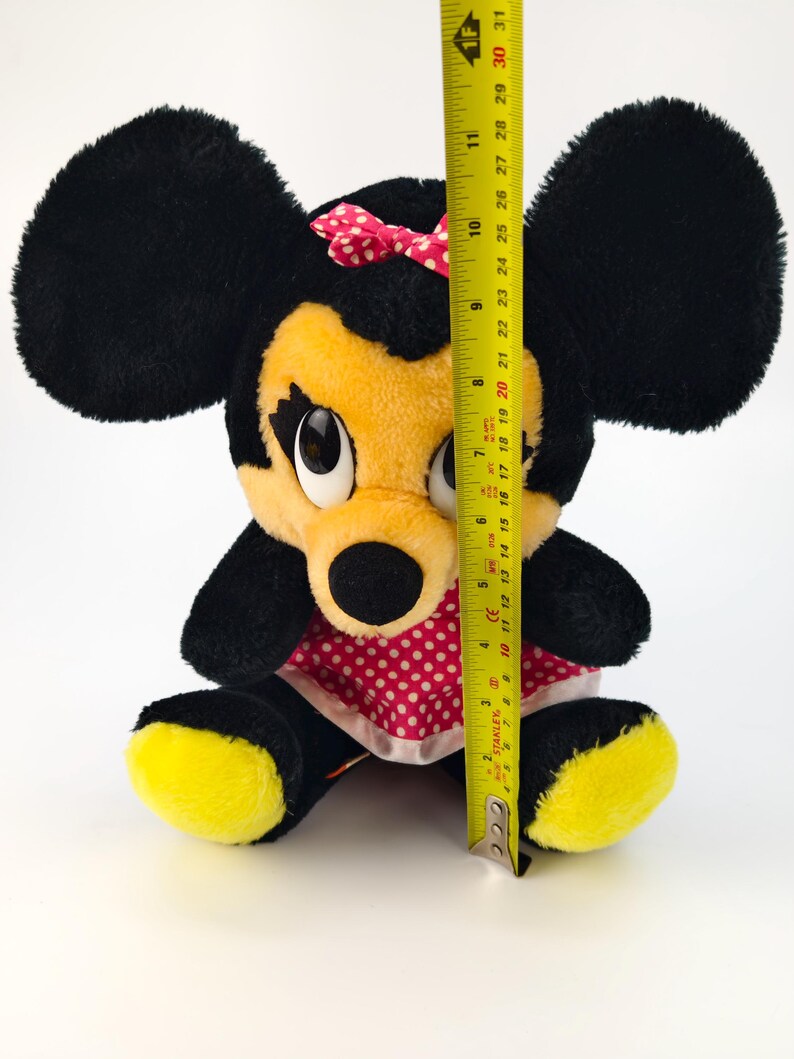 May include: A plush Minnie Mouse doll, featuring black fur, yellow face and feet, and a red polka-dot dress. The doll has a pink bow and is approximately 11 inches tall, as measured by a yellow tape measure.