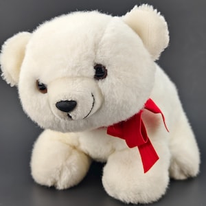 Puede incluir: Un oso de peluche blanco con un lazo rojo alrededor del cuello. El oso tiene ojos marrones y una nariz negra cosida. Está posado sobre una superficie gris oscuro.