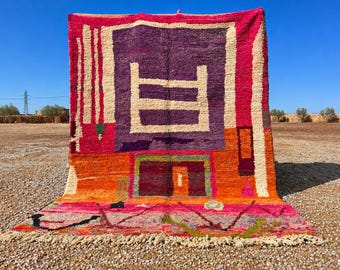 May include: A vibrant, hand-woven rug with a geometric design. The rug features a central purple square with a white rectangular pattern, surrounded by orange, pink, and cream colors. The rug is displayed outdoors.