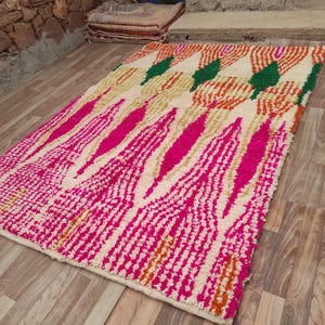 Puede incluir: Una alfombra rectangular con un dise&ntilde;o vibrante. La alfombra presenta un fondo crema con un patr&oacute;n de formas rosas, naranjas y verdes. La alfombra est&aacute; colocada sobre un suelo de madera, mostrando su textura y colores.