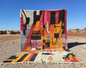 May include: A colorful, geometric patterned rug with a mix of red, black, yellow, orange, and white shapes. The rug is displayed outdoors against a clear blue sky, showcasing its vibrant design and texture. The rug's design includes abstract shapes and lines.