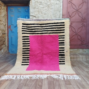 May include: A hand-woven rug with a geometric pattern. The rug is beige with black stripes and a large pink square in the center.