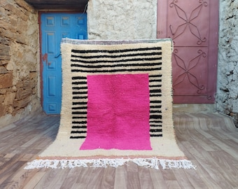 Könnte beinhalten: Ein handgewebter Teppich mit geometrischem Muster. Der Teppich ist beige mit schwarzen Streifen und einem großen pinkfarbenen Quadrat in der Mitte.