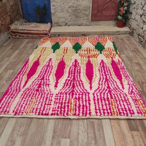 Puede incluir: Una alfombra rectangular con un dise&ntilde;o vibrante. La alfombra presenta un fondo color crema con un patr&oacute;n de formas geom&eacute;tricas rosas, naranjas y verdes. La alfombra est&aacute; colocada sobre un suelo de madera.