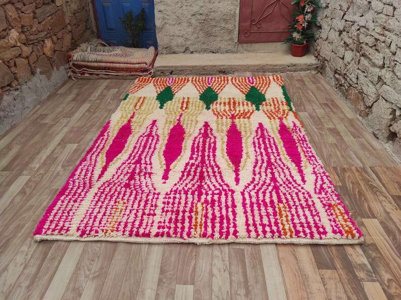 Puede incluir: Una alfombra rectangular con un dise&ntilde;o vibrante. La alfombra presenta un fondo color crema con un patr&oacute;n de formas geom&eacute;tricas rosas, naranjas y verdes. La alfombra est&aacute; colocada sobre un suelo de madera.