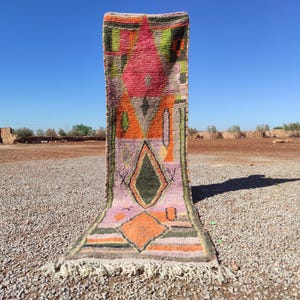 May include: A colorful Moroccan rug with geometric patterns in shades of pink, orange, green, and blue. The rug has a fringe border and is displayed against a backdrop of a desert landscape.