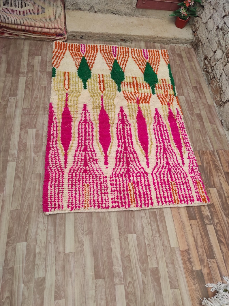 Puede incluir: Una alfombra rectangular con fondo crema y un patr&oacute;n geom&eacute;trico. El dise&ntilde;o presenta formas rosas, naranjas, verdes y doradas. La alfombra est&aacute; hecha de un material texturizado y est&aacute; colocada sobre un suelo de madera.
