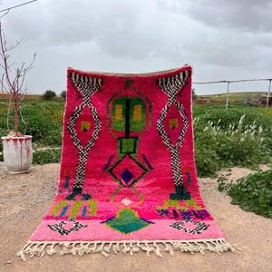 May include: A vibrant pink Moroccan rug with a geometric design. The rug features a central pattern with green, yellow, and blue accents, framed by black and white diamond shapes. The rug has a fringe along the bottom edge.