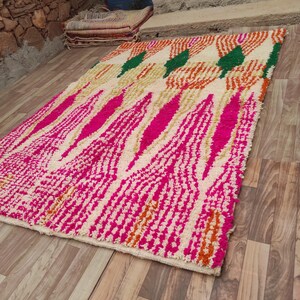 Puede incluir: Una alfombra rectangular con un fondo crema y un patr&oacute;n vibrante. El dise&ntilde;o presenta formas geom&eacute;tricas rosas, naranjas y verdes en negrita. La alfombra est&aacute; colocada sobre un suelo de madera, mostrando su textura y color.