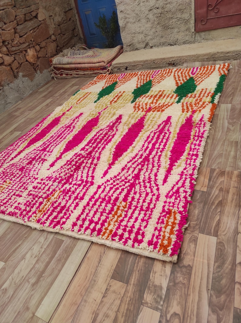 Puede incluir: Una alfombra rectangular con un fondo crema y un patr&oacute;n vibrante. El dise&ntilde;o presenta formas geom&eacute;tricas rosas, naranjas y verdes en negrita. La alfombra est&aacute; colocada sobre un suelo de madera, mostrando su textura y color.
