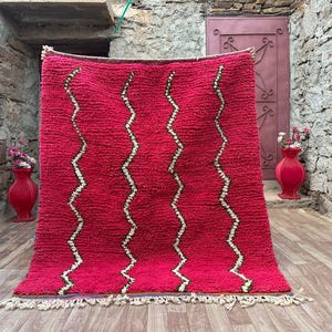 May include: A vibrant red rug with a textured surface and a zigzag pattern in white and beige. The rug has a fringe along the bottom edge. The rug is displayed in front of a stone wall.