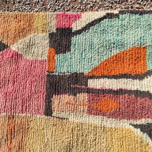 Op de afbeelding: Een kleurrijk abstract tapijt met geometrische vormen in tinten roze, oranje, blauw en bruin. Het tapijt is gemaakt van een dik, getextureerd materiaal.