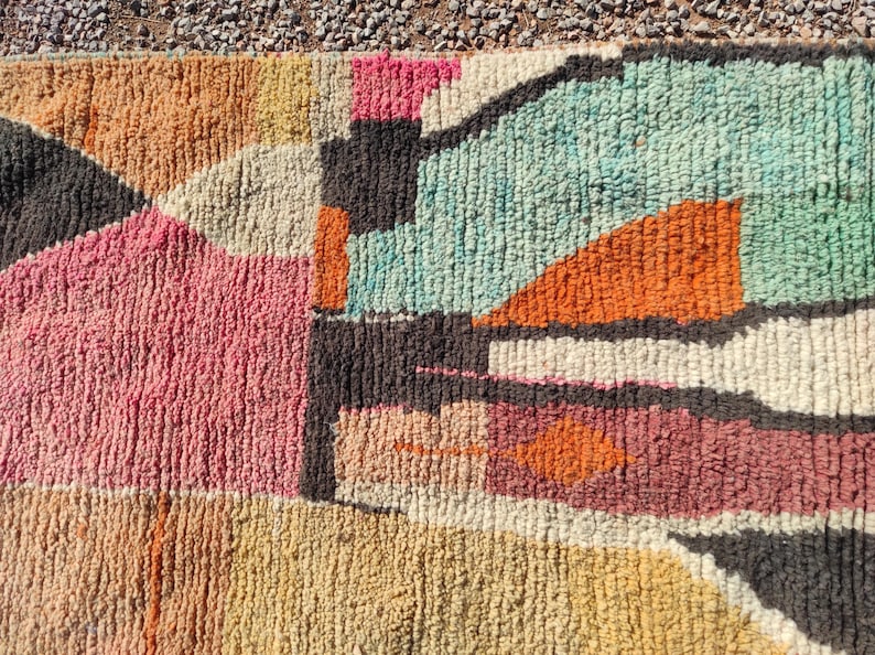 Op de afbeelding: Een kleurrijk abstract tapijt met geometrische vormen in tinten roze, oranje, blauw en bruin. Het tapijt is gemaakt van een dik, getextureerd materiaal.