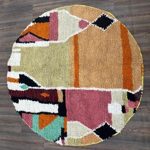 Może przedstawiać: Okrągły, teksturowany dywan z geometrycznym wzorem. Dywan ma różne kolory, w tym różowy, pomarańczowy, czarny i zielony, ułożone w abstrakcyjne kształty. Ma żółtą obwódkę i nadaje się do każdego pomieszczenia.