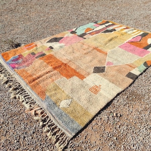 Op de afbeelding: Een kleurrijk, handgeweven tapijt met geometrische patronen in tinten van oranje, roze, geel, blauw en grijs. Het tapijt heeft een franje rand en is gemaakt van natuurlijke vezels.