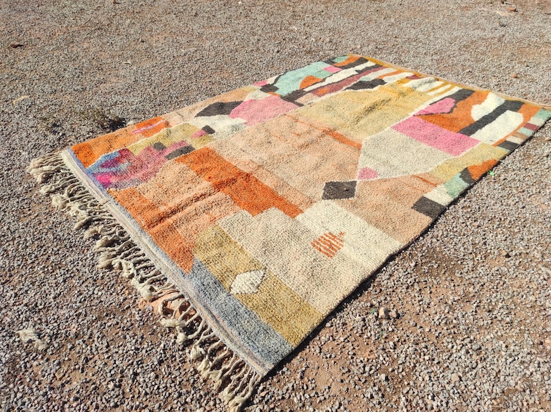 Op de afbeelding: Een kleurrijk, handgeweven tapijt met geometrische patronen in tinten van oranje, roze, geel, blauw en grijs. Het tapijt heeft een franje rand en is gemaakt van natuurlijke vezels.