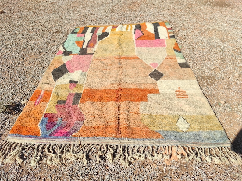 Op de afbeelding: Een kleurrijk Marokkaans tapijt met geometrische patronen in tinten roze, oranje, blauw en geel. Het tapijt heeft een franje rand en is gemaakt van wol.