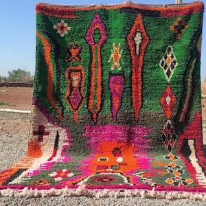 May include: A colorful Moroccan rug with a green background and intricate patterns in shades of pink, orange, and black. The rug has a fringe border and is woven with a high pile.