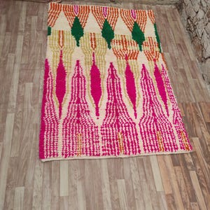 Puede incluir: Una alfombra rectangular con fondo crema y un patr&oacute;n geom&eacute;trico. El dise&ntilde;o presenta formas rosas, naranjas, verdes y doradas. La alfombra est&aacute; hecha de un material texturizado y est&aacute; colocada sobre un suelo de madera.