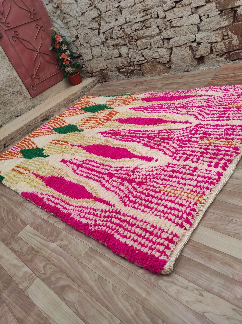 Puede incluir: Una alfombra rectangular con un dise&ntilde;o vibrante. La alfombra presenta un fondo color crema con un patr&oacute;n de formas rosa intenso, verdes y naranjas. La alfombra est&aacute; hecha de un material grueso y texturizado.