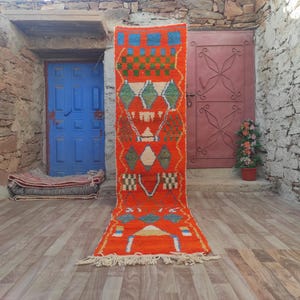 Pode incluir: Um tapete comprido e estreito, com padrão vermelho e azul, com franjas nas extremidades. O tapete está colocado sobre um piso de madeira na frente de uma porta azul e uma porta vermelha.