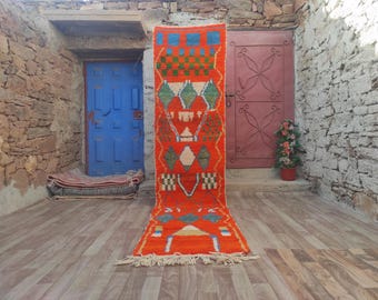 May include: A long, narrow, red and blue patterned rug with fringe on the ends. The rug is laid out on a wooden floor in front of a blue door and a red door.