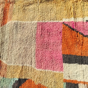 Op de afbeelding: Een close-up van een kleurrijk geweven tapijt met geometrische vormen in tinten oranje, roze, beige en grijs.