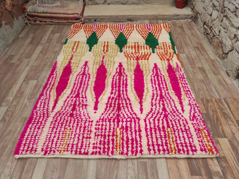Puede incluir: Una alfombra rectangular con un fondo crema y un patr&oacute;n vibrante. El dise&ntilde;o presenta formas verticales rosas y doradas, con formas geom&eacute;tricas naranjas y verdes en la parte superior. La alfombra est&aacute; colocada sobre un suelo de madera.