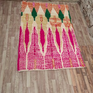 Puede incluir: Una alfombra rectangular con fondo crema y un patr&oacute;n geom&eacute;trico. El dise&ntilde;o presenta formas rosas, naranjas, verdes y doradas. La alfombra est&aacute; colocada sobre un suelo de madera.