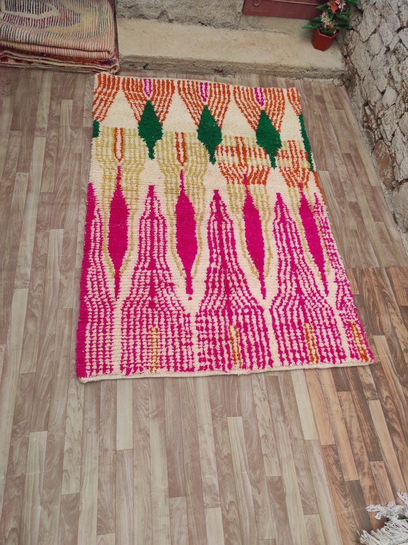 Puede incluir: Una alfombra rectangular con fondo crema y un patr&oacute;n geom&eacute;trico. El dise&ntilde;o presenta formas rosas, naranjas, verdes y doradas. La alfombra est&aacute; colocada sobre un suelo de madera.