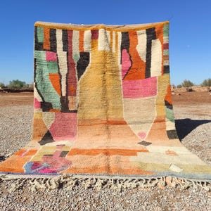Op de afbeelding: Een kleurrijk, abstract tapijt met geometrische patronen in tinten roze, oranje, geel, groen en bruin. Het tapijt heeft een franje rand en is gemaakt van wol.