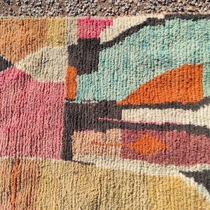 Op de afbeelding: Een kleurrijk abstract tapijt met geometrische vormen in tinten roze, oranje, bruin en blauw. Het tapijt is gemaakt van een dik, getextureerd materiaal.