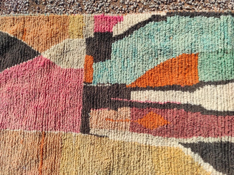 Op de afbeelding: Een kleurrijk abstract tapijt met geometrische vormen in tinten roze, oranje, bruin en blauw. Het tapijt is gemaakt van een dik, getextureerd materiaal.