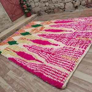 Puede incluir: Una alfombra rectangular con un dise&ntilde;o vibrante. La alfombra presenta un fondo color crema con un patr&oacute;n de formas rosa intenso, verdes y naranjas. La alfombra est&aacute; hecha de un material grueso y texturizado.