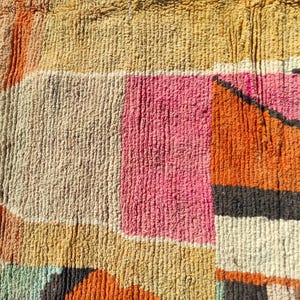 Op de afbeelding: Een close-up van een kleurrijk geweven tapijt met geometrische vormen in tinten oranje, roze, beige en grijs.