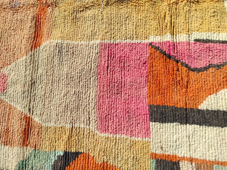 Op de afbeelding: Een close-up van een kleurrijk geweven tapijt met geometrische vormen in tinten oranje, roze, beige en grijs.