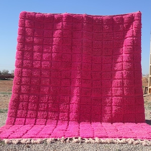May include: A pink hand-woven rug with a square pattern and fringe. The rug is made of wool and is approximately 5 feet by 7 feet.