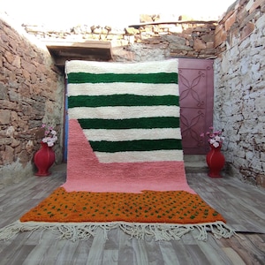 May include: A hand-woven rug with a geometric design featuring green, white, pink, and orange stripes. The rug has a fringe border and is displayed against a stone wall.