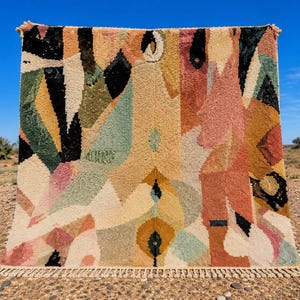 Peut inclure: Un tapis rectangulaire à poils longs présentant un motif géométrique abstrait. Le design intègre une palette de couleurs crème, noir, vert sauge, jaune moutarde et vieux rose, avec une finition à franges nouées sur le bord inférieur.
