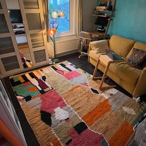 May include: A colourful, abstract patterned rug with a mix of pink, orange, green, and brown hues. The rug is laid out on a dark wood floor in a living room setting.