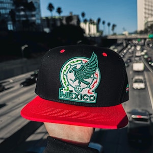Op de afbeelding: Zwarte en rode baseballcap met het Mexicaanse wapen. De cap heeft een platte rode klep en een zwarte kroon met een groen en wit embleem en het woord "MEXICO".