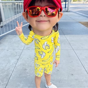 Op de afbeelding: Een kind draagt een gele outfit met bananenprint, een rode zonnebril, een rode pet met een ananasontwerp en witte klompen. Het kind maakt het vredesteken.