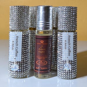 May include: Several perfume bottles with silver caps and labels that read "French Coffee" and "Al-Rehab." The central bottle has a brown liquid and a label that says "Crown Perfumes" and "French Coffee Concentrated Perfume." The bottles are decorated with rhinestones.