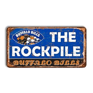 Puede incluir: Un letrero rectangular de metal con un acabado azul desgastado que presenta el logotipo de los Buffalo Bills, el texto THE ROCKPILE en letras blancas grandes y el texto BUFFALO BILLS en naranja en la parte inferior, con un borde marrón oxidado.