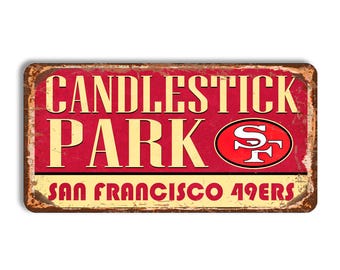 Puede incluir: Un letrero rectangular de metal con acabado desgastado que presenta el texto CANDLESTICK PARK en letras grandes de color crema sobre un fondo rojo. El logo de los San Francisco 49ers está a la derecha y SAN FRANCISCO 49ERS en rojo abajo.
