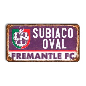 May include: A rectangular metal sign with a distressed, vintage appearance. The sign displays the words "SUBIACO OVAL" in white, with "FREMANTLE FC" below. A logo with a man holding an anchor is on the left.