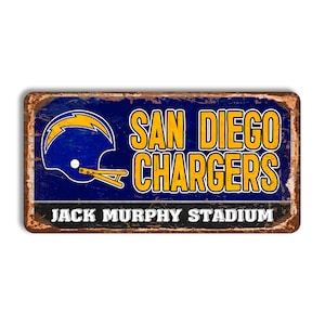 Puede incluir: Un letrero de metal de estilo vintage con un acabado oxidado. Presenta el logotipo de los San Diego Chargers, un casco azul con un rayo, y el texto "San Diego Chargers Jack Murphy Stadium".