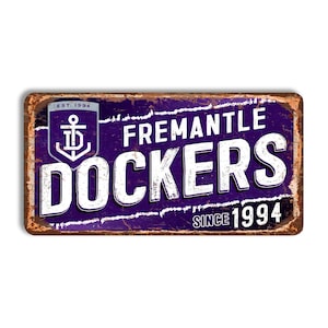 May include: A rectangular metal sign with a distressed finish. The sign features the words "FREMANTLE DOCKERS" in white block letters on a purple background. An anchor logo is in the upper left corner, with the text "EST. 1994".