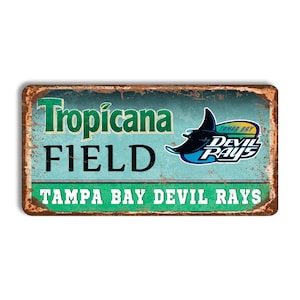 May include: A vintage-style metal sign with a rusty finish. The sign features the text "Tropicana Field" in green and black lettering. The Tampa Bay Devil Rays logo is featured on the right side of the sign. The sign also includes the text "Tampa Bay Devil Rays" in green lettering.