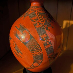 May include: A striking orange ceramic vase with a bulbous form and a slender neck. The vase is adorned with detailed black geometric designs that encircle its exterior. It rests on a black base, displaying a fusion of traditional craftsmanship and contemporary style.